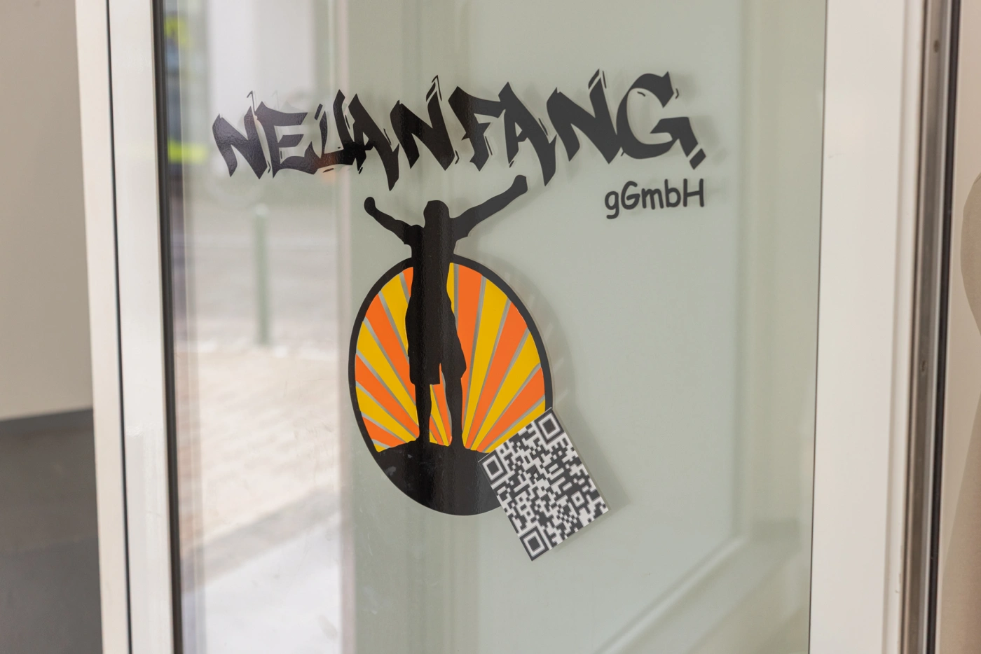 Logo der Neuanfang gGmbH mit einer stilisierten Figur und QR-Code auf einer Glastür.