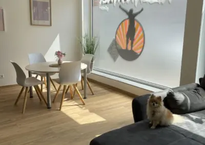 Ein Wohnraum mit einem runden Tisch, Stühlen und einem Sofa, sowie einem Fenster mit einem grafischen Element.
