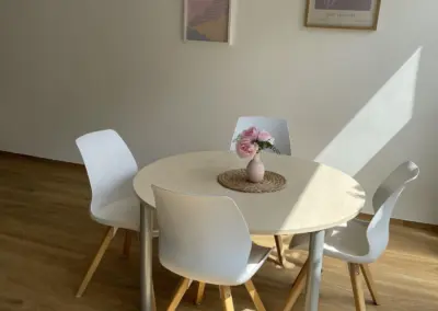 Runder Tisch mit vier weißen Stühlen und einer Vase mit Blumen auf einem Tischläufer.