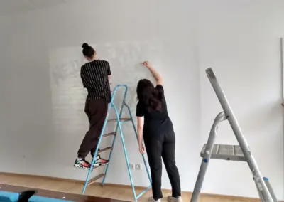 Zwei Personen, eine auf einer Leiter und die andere am Boden, malen an einer weißen Wand.