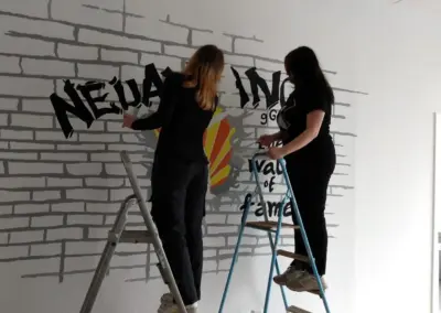 Zwei Frauen stehen auf Leitern und malen ein Wandgemälde mit schwarzer Schrift auf einer weißen Ziegelwand.