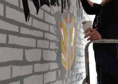 Frau steht auf einer Leiter und malt ein Graffiti-Wandgemälde mit dem Wort 'Neuanfang' und einem Wappen.