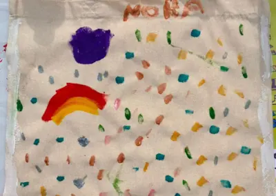 Stofftasche mit bunten Farbflecken und einem Regenbogen sowie dem Namen 'Nora' darauf.