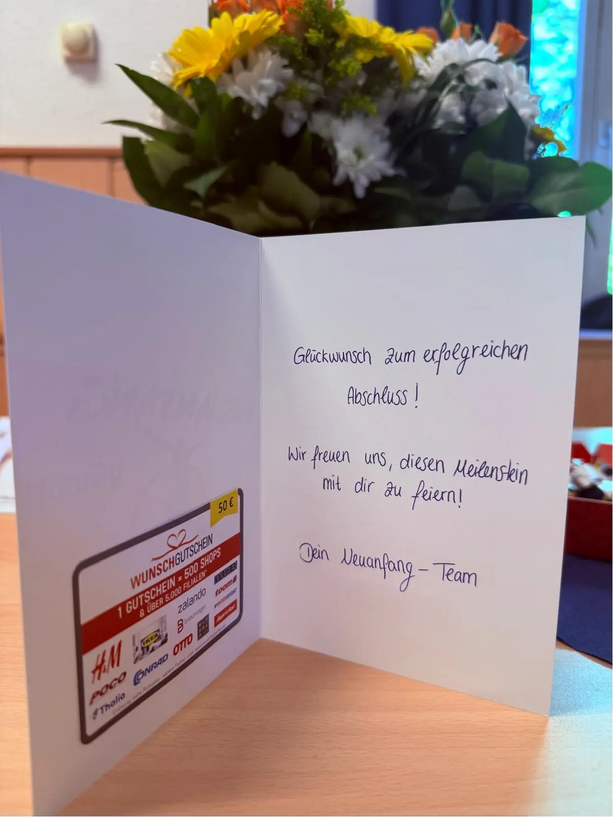 erfolgreiche-abschluesse_3 Eine Glückwunschkarte mit einem Gutschein und Blumenarrangement im Hintergrund.