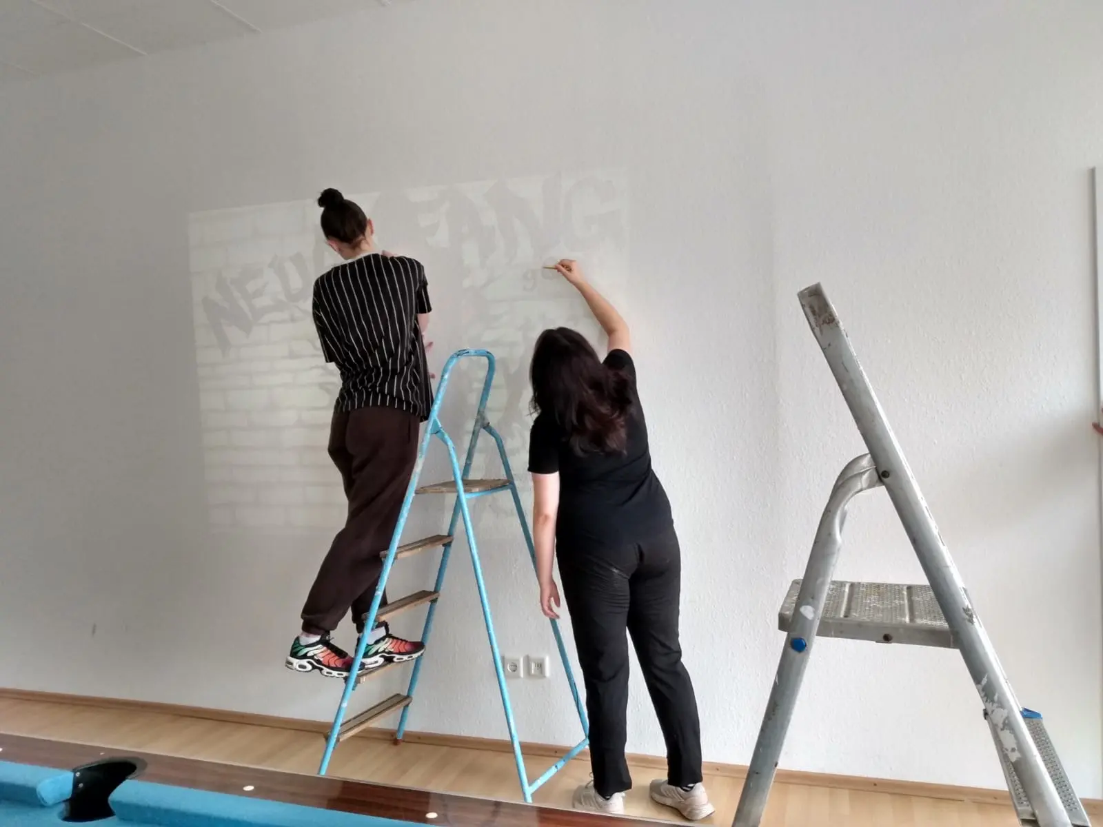 Zwei Personen, eine auf einer Leiter, die eine Wand mit einem Graffiti-Motiv bemalen.