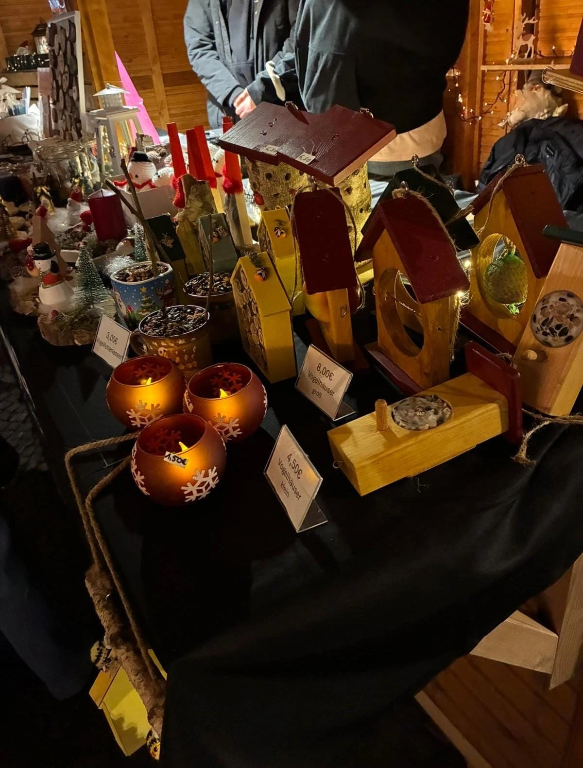 neuanfang-hamm_weihnachtsmarkt-2025-1 Bunte VogelhÀuser und dekorative Kerzen auf einem Tisch, prÀsentiert auf einem Weihnachtsmarkt mit verschiedenen handgefertigten Artikeln.