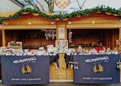 Holzhütte auf einem Weihnachtsmarkt mit dekorierten Ständen und verschiedenen handgefertigten Artikeln. Banner mit dem Logo von Neuanfang Hamm.