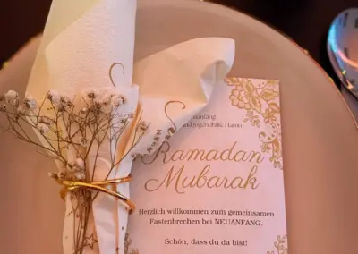 Tischdekoration mit einer Serviette, die mit einem goldenen Band und Trockenblumen gebunden ist, sowie einer Karte mit der Aufschrift 'Ramadan Mubarak'.