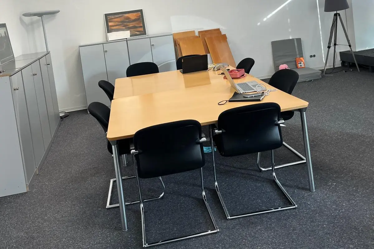 Warstein Betreuungsbüro Meetingraum mit holzfarbenem Tisch, schwarzen Stühlen, Laptop, Ordnern und Ablagen, ergänzt durch moderne Beleuchtung und Laminatboden