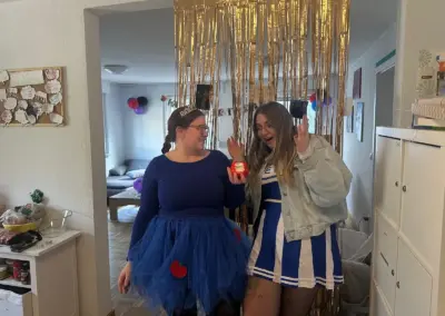 Zwei junge Frauen bei einer Geburtstagsfeier in Innenräumen, blaues Kostüm mit Tüllrock und Tiara neben einer Frau in blau-weißer Kleid-Uniform; goldene dekorative Hintergrundgirlanden hängen hinter ihnen.