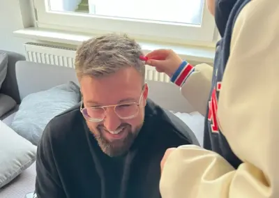 Mann mit grauen Haaren und runder Brille sitzt lächelnd auf einer Couch, während eine weitere Person ihm die Haare stylt, im hellen Wohnzimmer mit Fenster im Hintergrund.