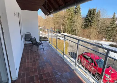 Balkon mit Fliesenboden und zwei Stühlen, Blick auf eine rote Auto und Bäume im Hintergrund.