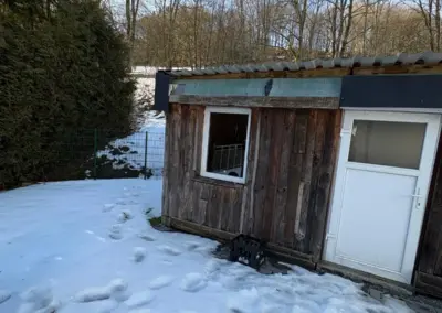 Holzschuppen mit einer weißen Tür und einem schneebedeckten Boden in einer winterlichen Umgebung.