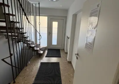 Eingangsbereich mit Treppe, zwei Matten auf dem Boden und weißen Wänden in einem Gebäude.