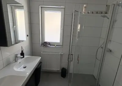 Modernes Badezimmer mit Dusche, Waschbecken und Spiegel. Weiße Fliesen und graue Bodenfliesen sind vorhanden.