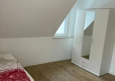 Schlafzimmer mit einem Einzelbett, einer weißen Kommode mit Spiegel und einem Fenster unter einer schrägen Decke.