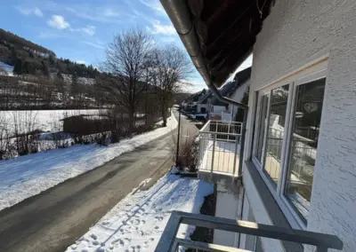 Balkonansicht eines Ferienhauses in Winterberg mit schneebedecktem Boden und klarer Himmel. Die Straße verläuft entlang des Hauses.