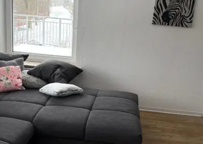 Dunkelgraues Sofa mit Kissen und einem kleinen Tisch im Vordergrund, Fenster mit Blick auf die Außenansicht.