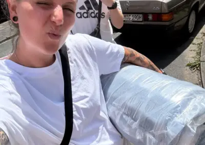 Zwei junge Erwachsene stehen in der Sonne vor einem älteren Mercedes und tragen eine in Folie verpackte Rolle, während einer ein Peace-Zeichen zeigt.