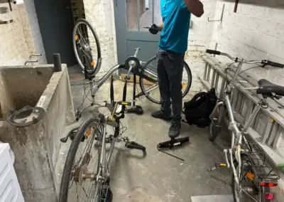 Eine Person im blauen Shirt repariert in einem Kellerraum ein umgedrehtes Fahrrad, während weitere Räder und Werkzeug um sie herum liegen.