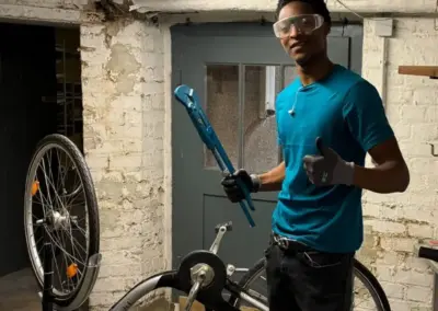Ein Mann in Schutzbrille und Handschuhen repariert in einem Kellerraum ein Fahrrad an der Werkbank, während eine Lampe den Raum erhellt.