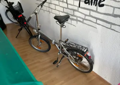 Zwei zusammengeklappte Fahrräder stehen in einem Raum vor einer weiß-grauen Ziegelwand mit dem Schriftzug „Wall of Fame“.