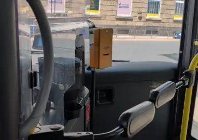 Im Bus sind Fahrgastraum, gelbe Haltestange und ein Entwerter zu sehen; draußen steht ein Gebäude mit Postbank-Schildern an der Straße im Sonnenlicht.