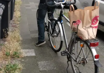 Ein Mann schiebt ein Fahrrad mit Einkaufstaschen neben einem Neuanfang-Hamm-Transporter in einer schmalen Wohnstraße zwischen Mehrfamilienhäusern.