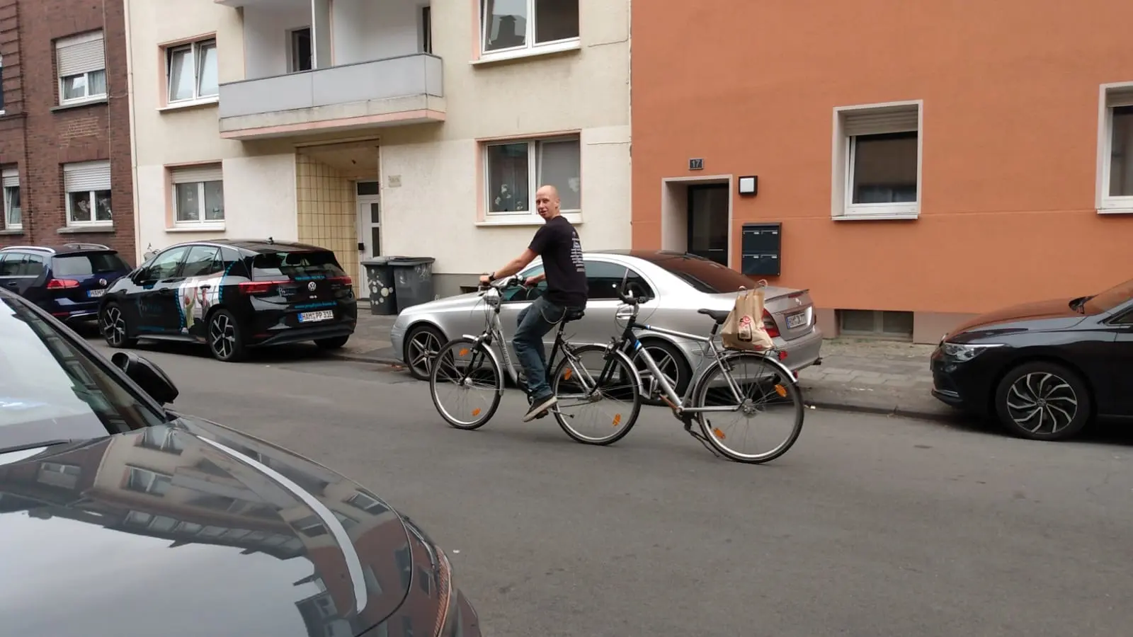 Ein Mann fährt auf einem Tandemfahrrad durch eine enge Wohnstraße mit geparkten Autos und Mehrfamilienhäusern während des autofreien Monats.